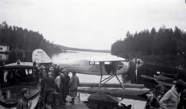 Historic photo, float plane at a dock with several people loading or unloading goods in the foreground, open water and trees in back landscape