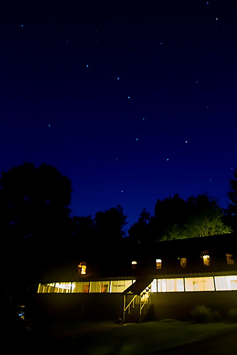 Stars shine over a large, brightly-lit building, with trees silhouetted on the horizon.