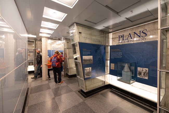 The interior of the exhibit level of the Washington Monument with a pole embedded from the ceiling to the ground in the background. An exhibit, "Plans for a Monument" has a glass case containing a miniature version of the Monument and sits to the right.