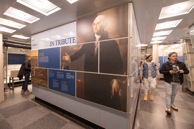 A wall exhibit "IN TRIBUTE" sits in the middle of the exhibit level of the Washington Monument as visitors roam the narrow pathways surrounding it.