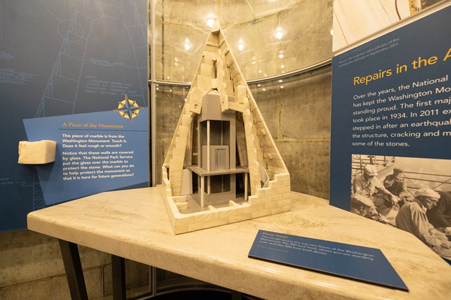 A miniature model of the top two floors of the Monument sits on a table in the foreground and a tactile exhibit of a marble piece of the monument is in the background.