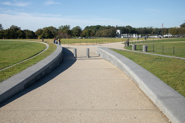 A paved path with curbing on either side and two bollards embedded in the ground in the middle of the path leading to the Washington Monument.