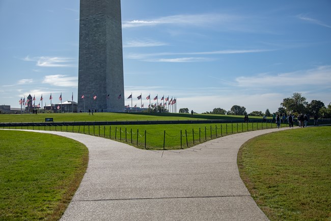 A paved, diverging path leads to the Washington Monument. The base of the Monument sits in the background surrounded by American flags in a circular pattern.