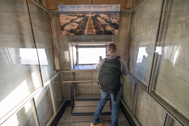 A visitor takes a photo of the outward view in one of the eight window areas on the observation level that has two steps and a handrail in front of the window.