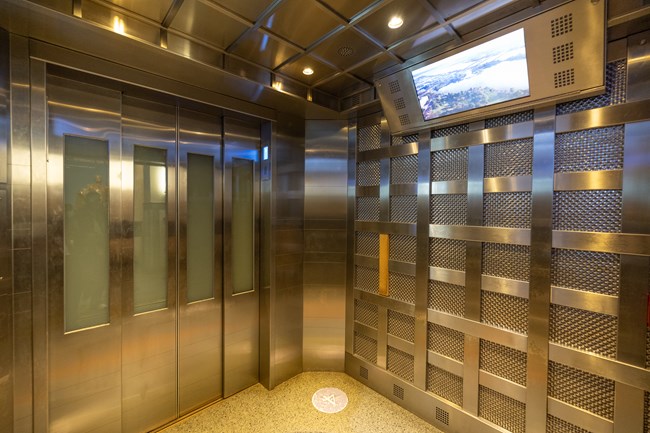 The inside of the Washington Monument elevator. The interior material is metallic and a TV sits atop the right wall.