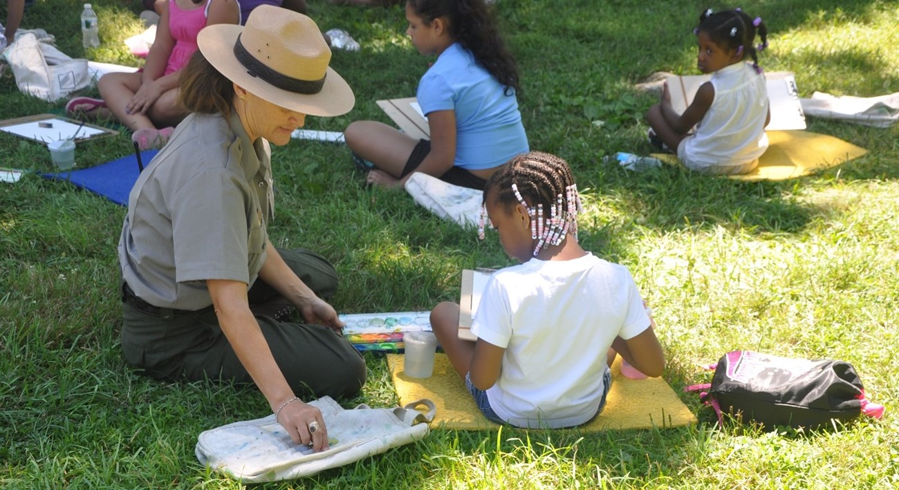 A women wearing a flat hat and a grey NPS shirt sits next to a girl painting.