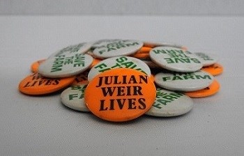 A pile of pins, some are orange that say Julian Weir Lives while some are white that say Safe the Farm.