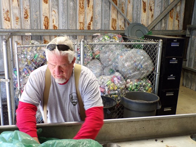 A caucasian man with white hair sorting recyclable items. Several bags of aluminum cans behind him,