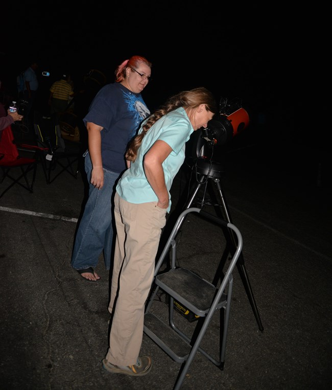 At nighttime, two visitors, one looking through the lens of a telescope.
