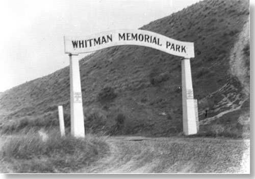 Historic photo of arch over dirt road. Text on arch say, "Whitman Memorial Park."