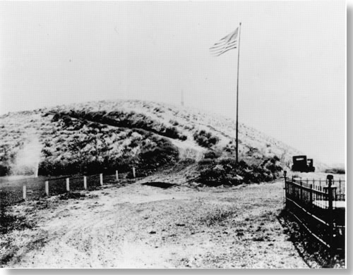 Historic photo of road leading up to hill. Large flagpole with American flag at base of hill. Cars parked near flagpole.