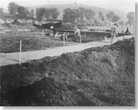 Stakes and ropes define archeological dig area. Three men working at site.