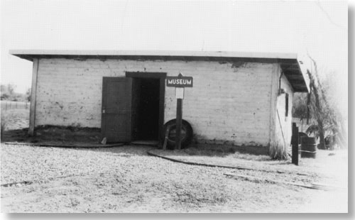 Historic photo of small, one story building. No windows on front wall. Wooden sign says