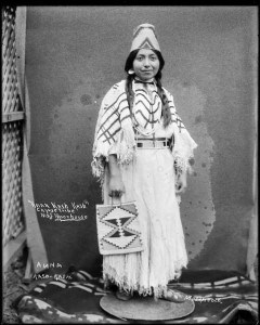 A black and white photo of a Native woman dressed in regalia looks towards the camera smiling. She is holding a beaded bag in one hand. The words "Anna Kash Kash" Cayuse Tribe Maj. Moorhouse are written on the photograph to her left.