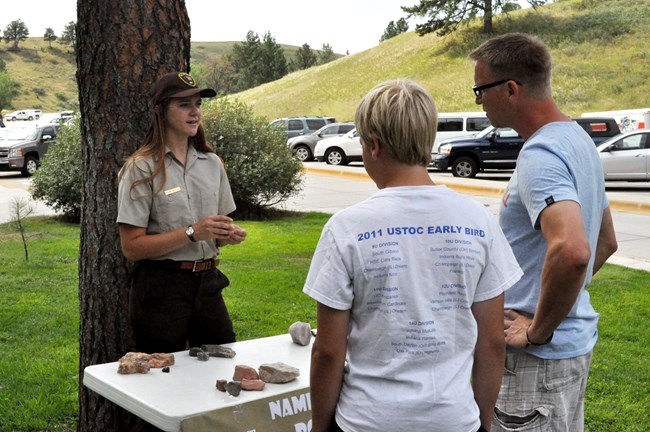 An intern stands at a table conducting a rock demonstration for two park visitors.