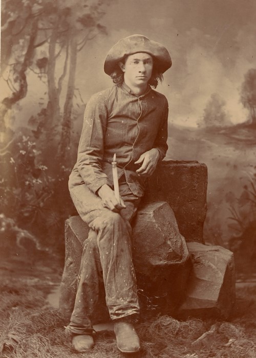 a sepia photo of a young man with a wide brimmed hat sitting on a rock holding a candle