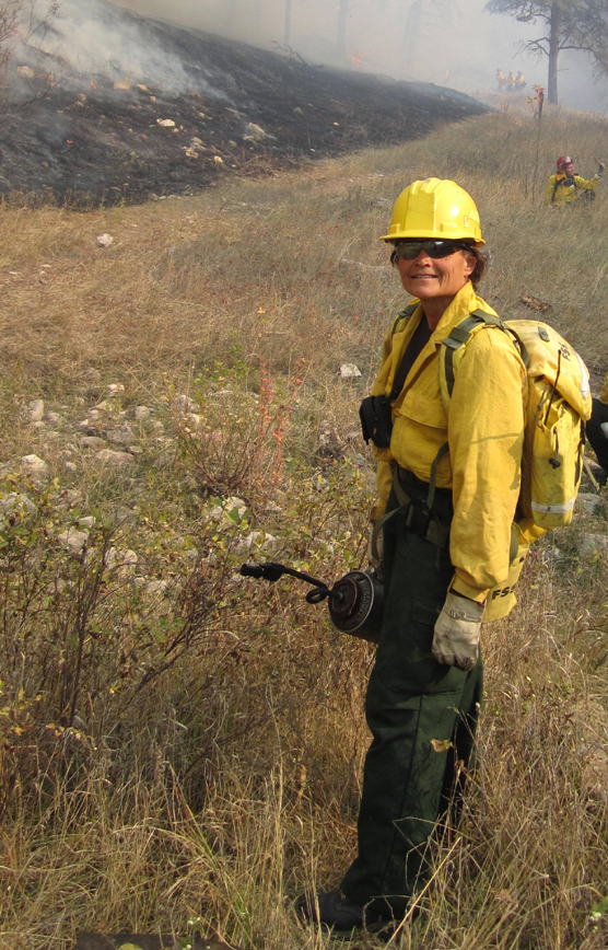 Beth Burkhart, dressed in yellow Nomex, standing near a fire line with a drip torch in her hand.
