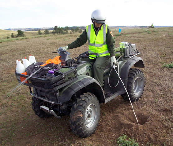 Biological technician Barb Muenchau sites on an ATV and uses a Techni-Duster to apply Deltamethrin into a prairie dog burrow.