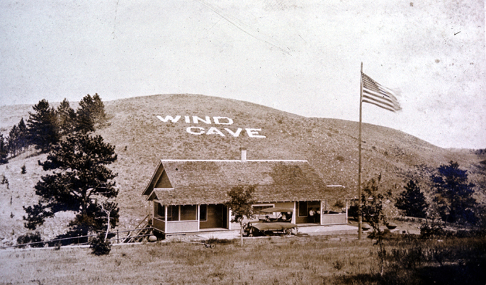 First park visitor center, 1920s era, with Wind Cave spelled out in white rocks on hillside behind visitor center.