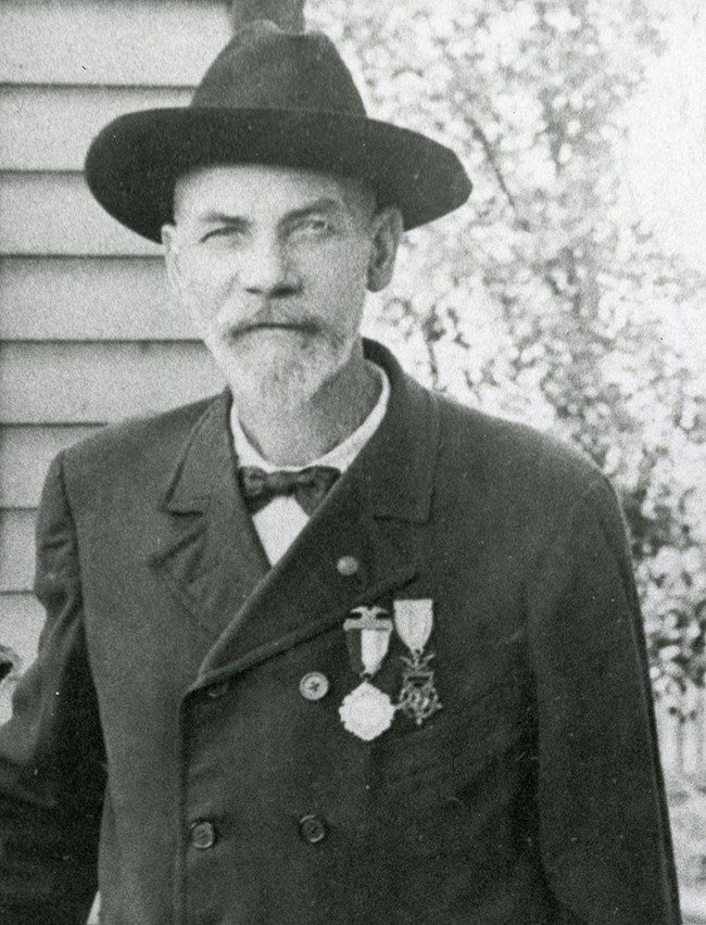 An older man in a civilian jacket and hat wears several military ribbons on his jacket