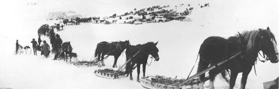 Historic photo of miners travelling in snow covered Thompson Pass with horses, sleds, and early bicycles.