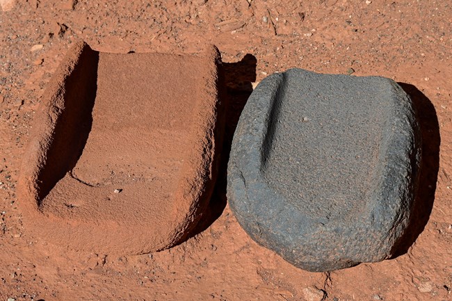 Two metates, or stone grinding tools, sit on red sand.