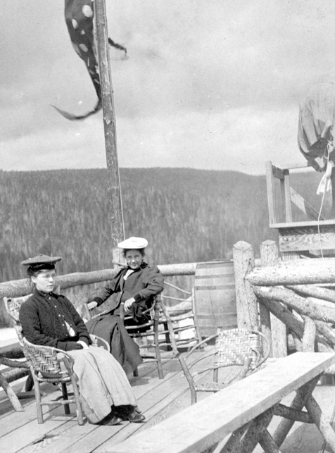 # YELL 118409. From 1906, Mrs. Albert Noyes and Adilade Bostick sitting on the widow’s walk of the Old Faithful Inn, the searchlight is on the right side of the image. 