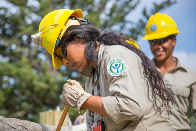 Native American youth digging, wearing a yellow hard hat. shoulder patch displays the Yellowstone Conservation Corps logo.