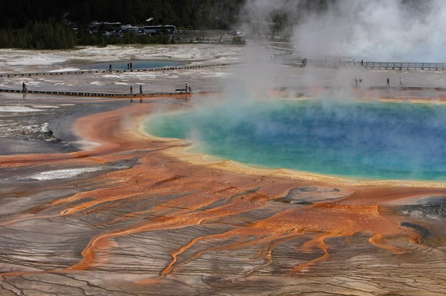 The rich oranges, yellows, and blues of a steaming hot spring with visitors walking next to it on a long boardwalk.