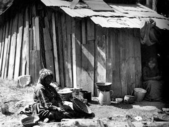 Callipene & Lena Brown sitting outside a cabin. Lena Brown sits by the doorway smiling as Callipene appears to be sitting among her baskets while taking a break from basket weaving.
