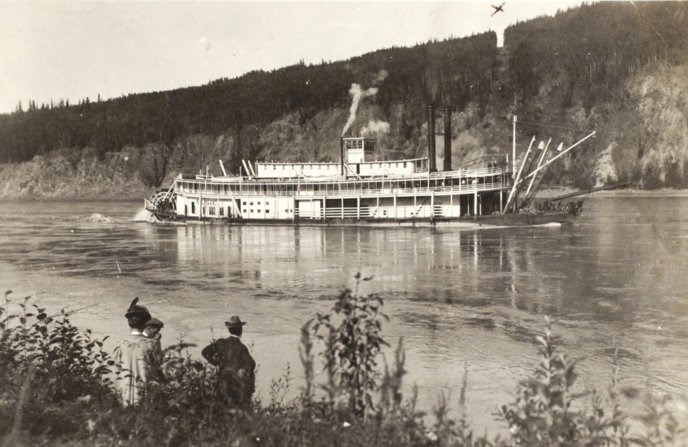 Historic photo of a riverboat on the Yukon River passing in front of the new international boundary which is a bare swath cut through the trees.
