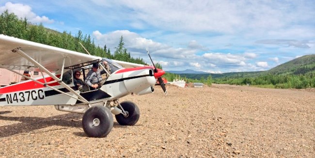Pilot & passenger sit inside a small bushplane parked at the Coal Creek Camp airstrip