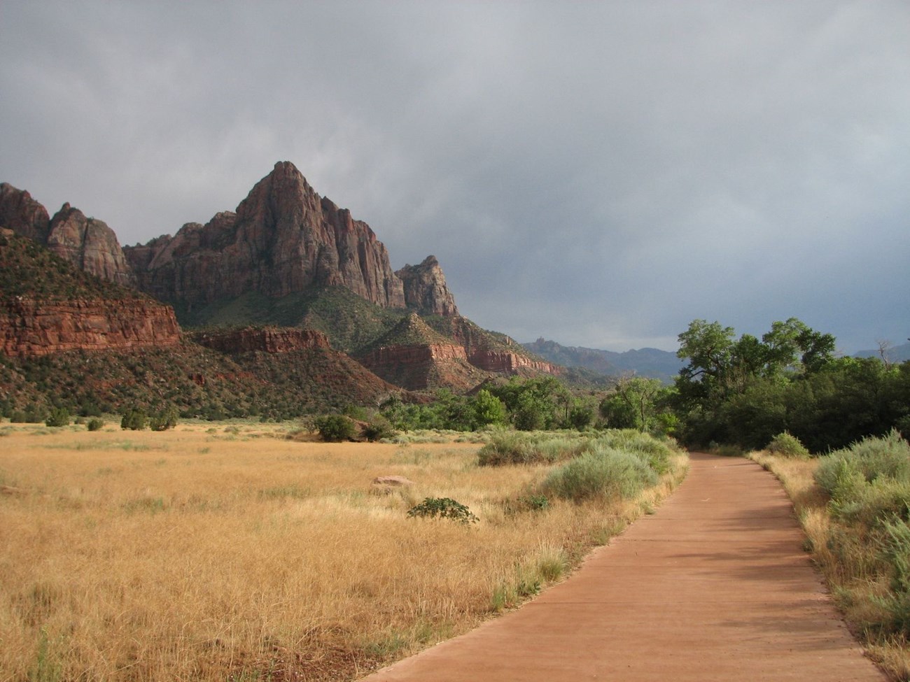 A paved path meanders through a desert scrubland. Watchman Tower, a 3,000 foot sandstone tower, is pictured in the background, with dark skies looming overhead