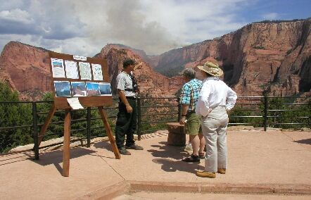 A ranger talks to a few visitors next to a board of information with smoke from a fire in Kolob Canyons behind them.