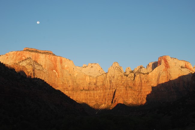 Warm sunlight illuminates orange cliffs, with a blue sky and a full moon.