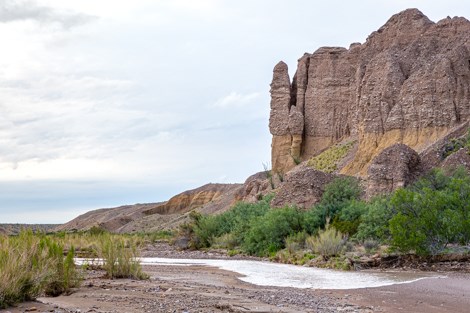 A line of water begins to fill a dry creek bed located next to a high cliff face.