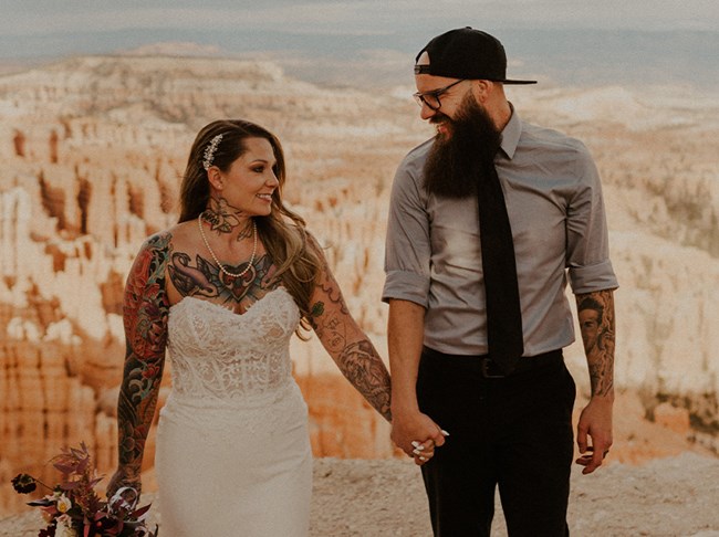 A man and woman holding hands, the man is staring at the woman who is wearing a wedding dress.