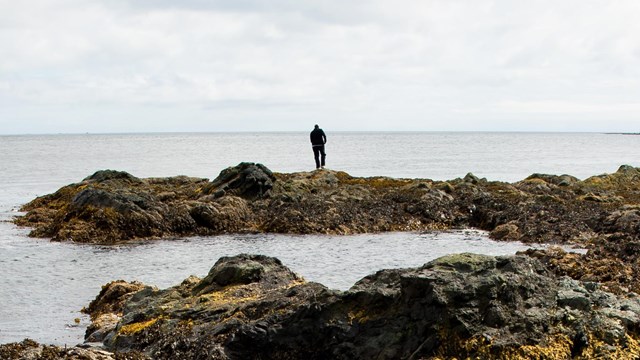 a person in the distance stands on a rocky spit overlooking a coastal bay.