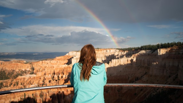 A woman with red hair, wearing a turquoise shirt looks out over the view with a rainbow above her.