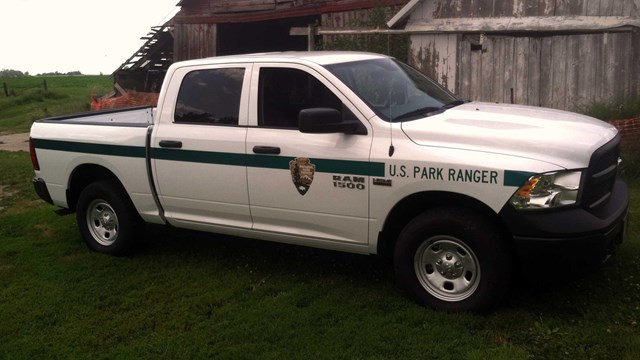 A park ranger patrol truck, painted white with a green stripe, is parked by a barn.