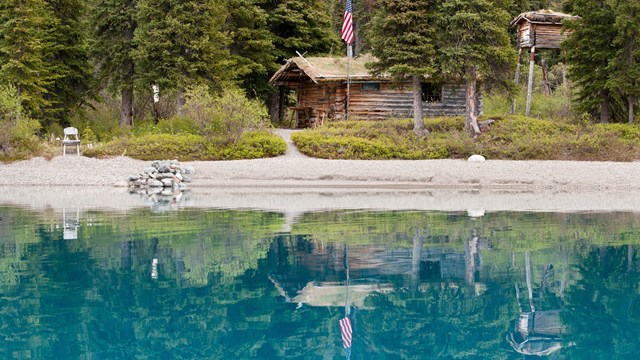 Photograph of a small log cabin nestled in the woods reflecting in a blue-green lake.