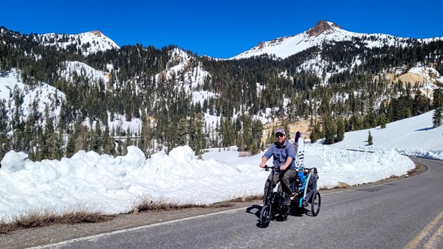 A man rides a small bicycle pulling a trailer on a road lined by snow.