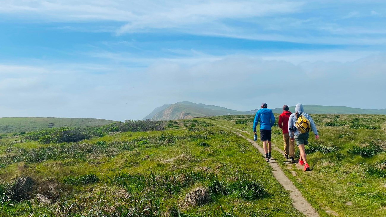 Three people hike along a dirt path through coastal grasslands under a blue partly cloudy sky.