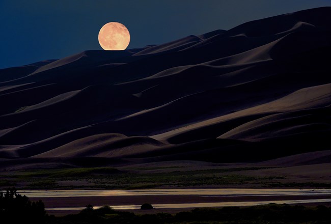 The full moon is over tall dunes and reflected in a shallow stream below