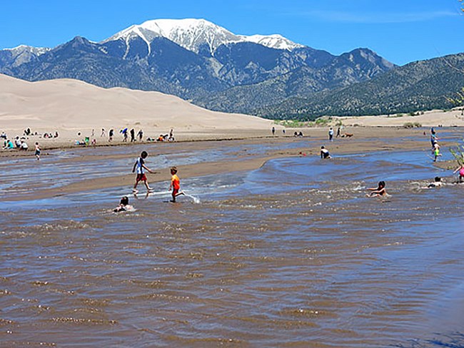 A wide shallow creek flowing at the base of dunes and a snowy mountain with children playing