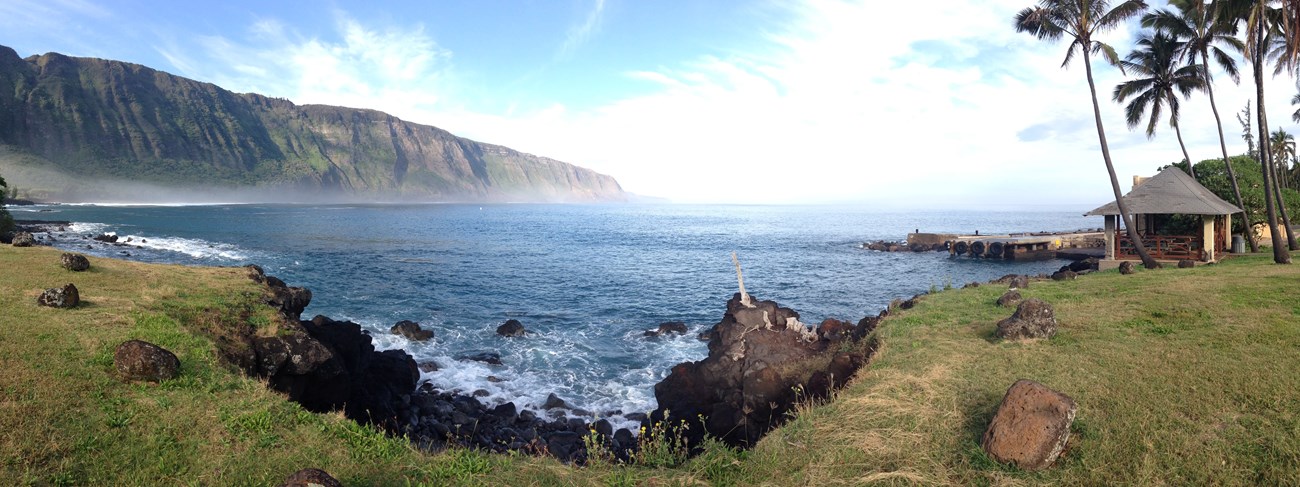 A panorama image of tall sea cliffs on the ocean on the left side, on the right side a pier, shade structure and palm trees stand next to the ocean.