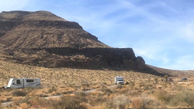 A bluff in the background with RVs. A campfire pit and picnic table in the foreground. 