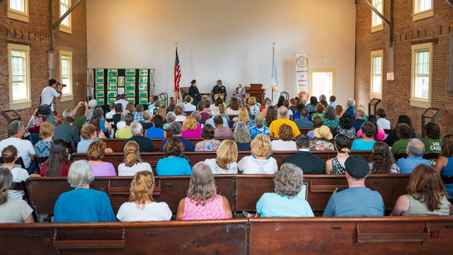 A crowd sitting in rows of wooden pews in the Wesleyan Chapel, watching a living history performance