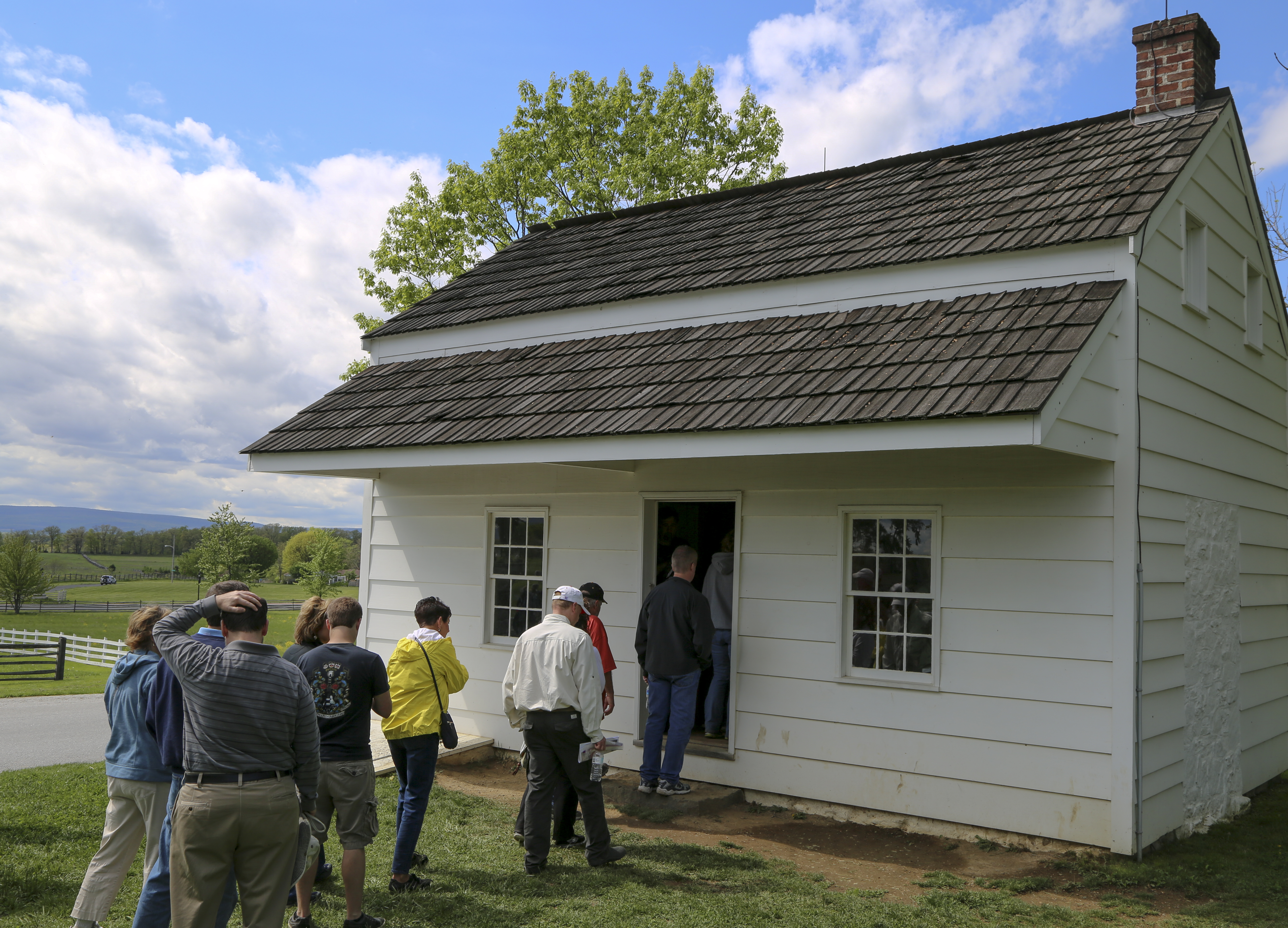 A group of visitors wait in line to get inside a small white sided historic house. The doorway into the house is in the middle and small windows flank the door on both sides. There is a red brick chimney on the upper right of the roof.