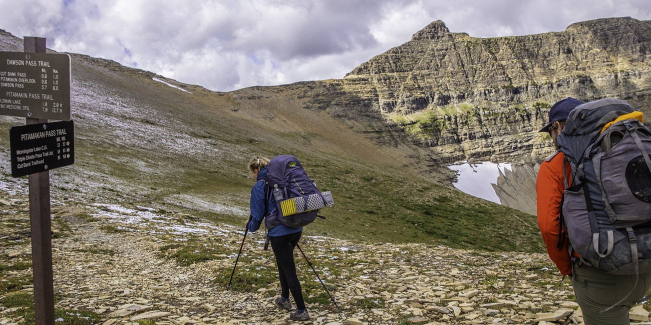 Two backpackers pass a trail junction sign on a ridgeline.